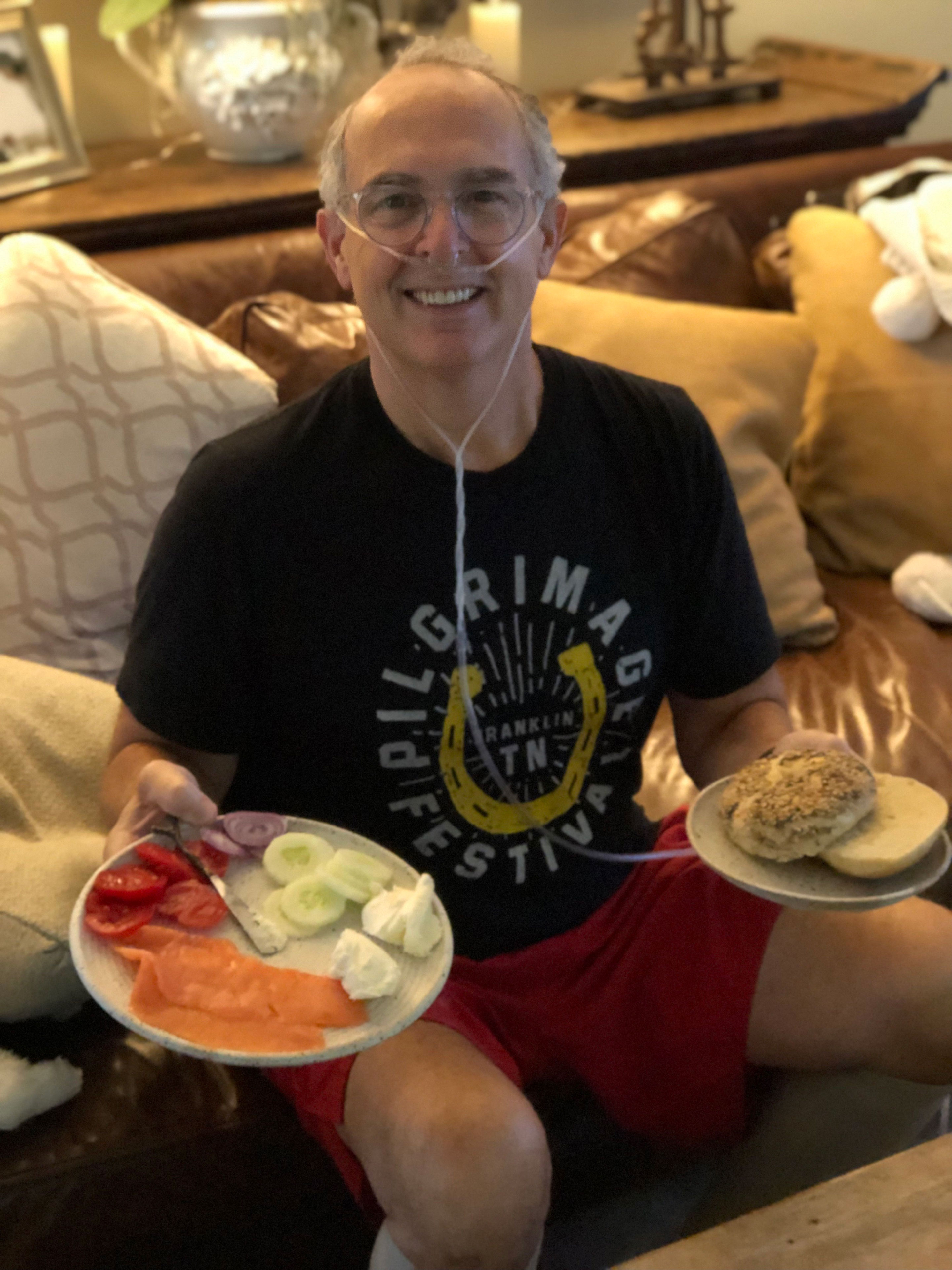 A smiling man wearing a nasal cannula sits on a couch holding a plate of food in each hand. 