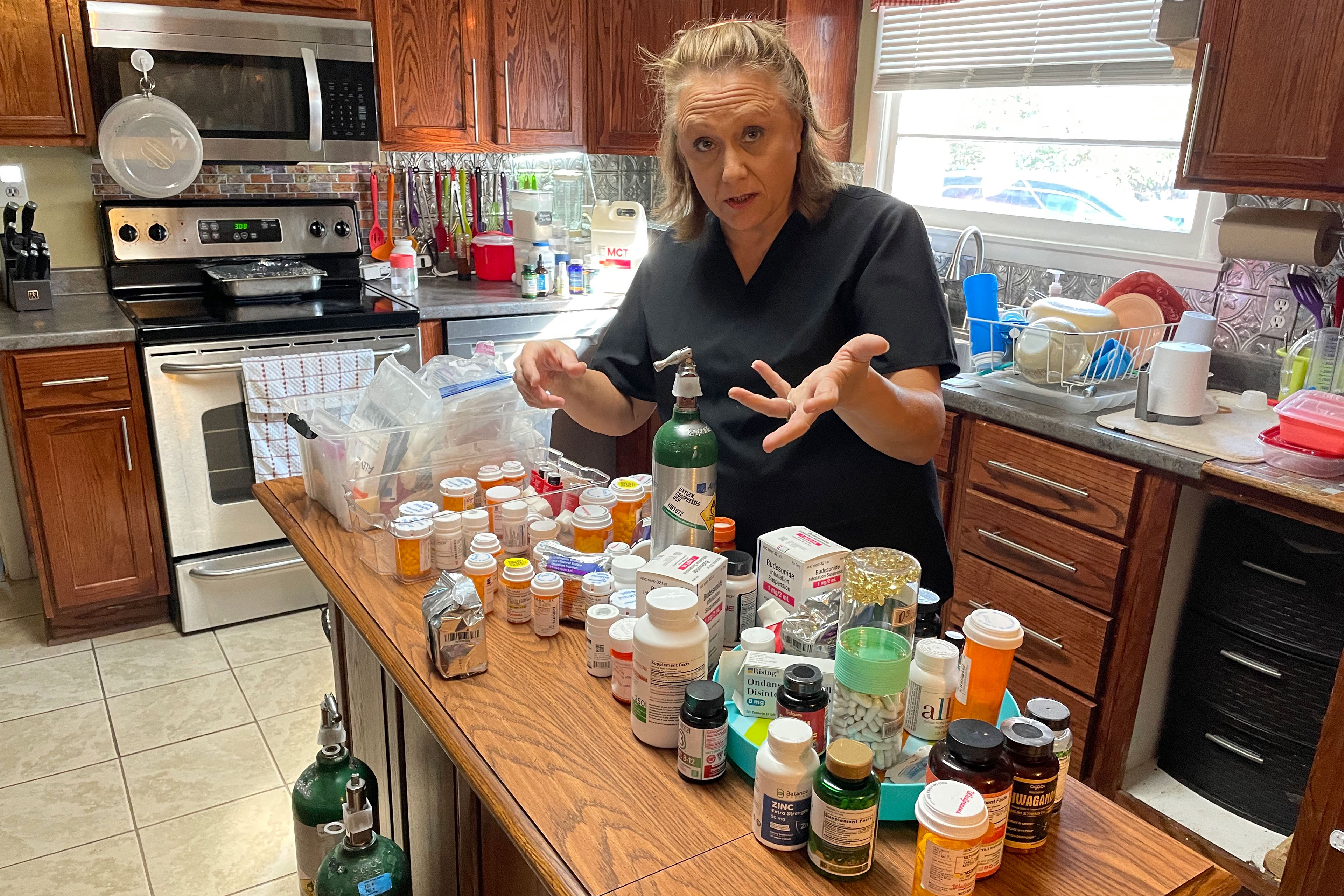 A woman stands at a kitchen island where the counter is filled with dozens of medication bottles. 