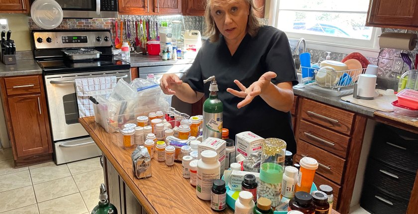 A woman stands at a kitchen island where the counter is filled with dozens of medication bottles.