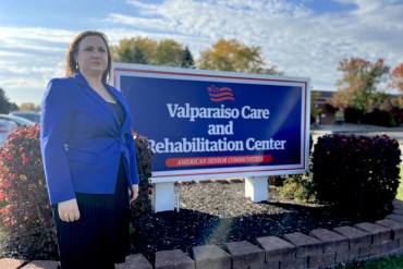 A photo shows Susie Talevski standing next to the sign outside the Valparaiso Care and Rehabilitation Center, the nursing home where her father resided.