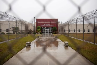 A photo shows the front entrance of Stewart Detention Center through a chain-link gate.