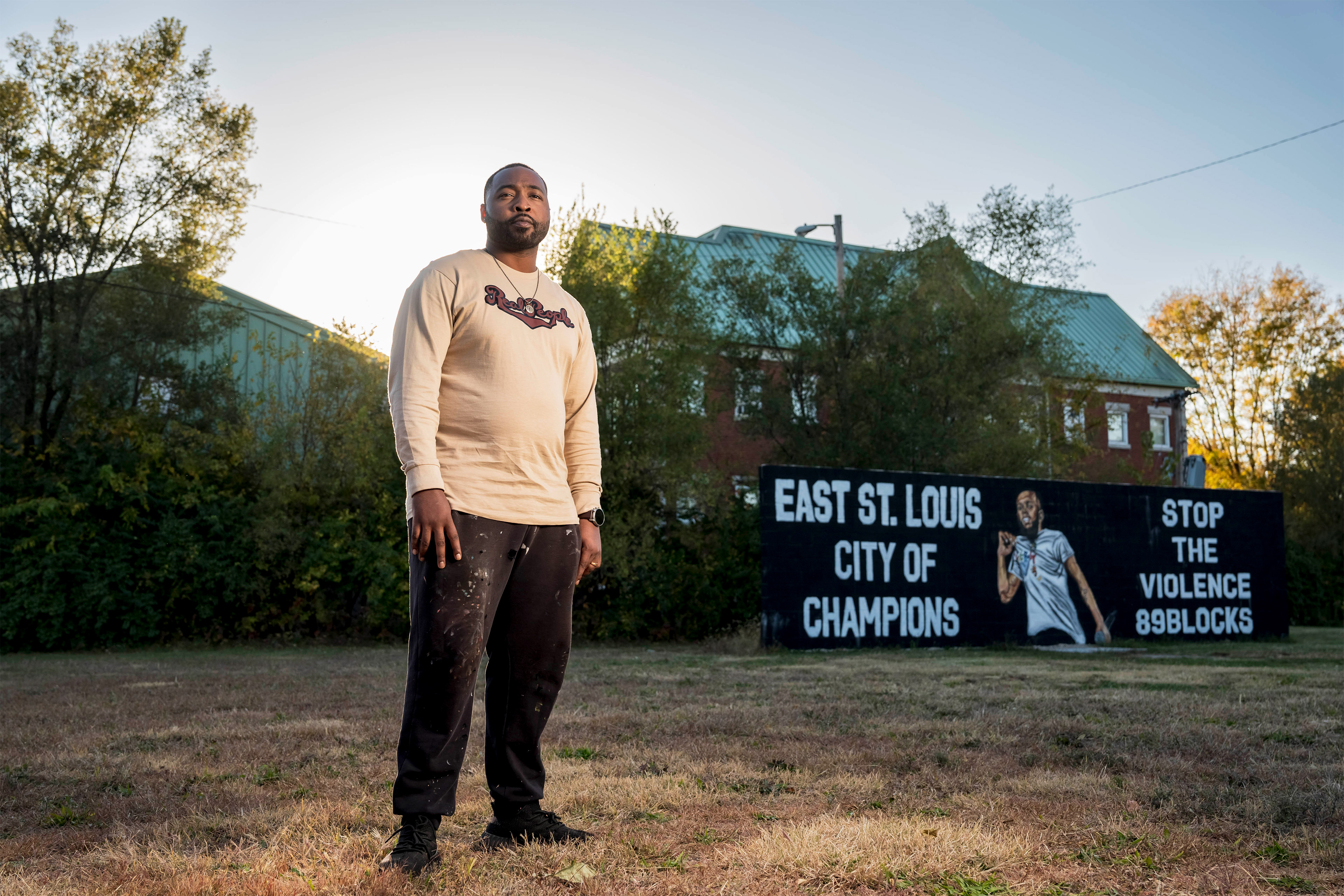 A photo shows Mykael Ash standing outside by a mural he painted of rapper Cedric Gooden. Text on the mural reads, "East St. Louis City of Champions" and "Stop the violence. 89blocks."