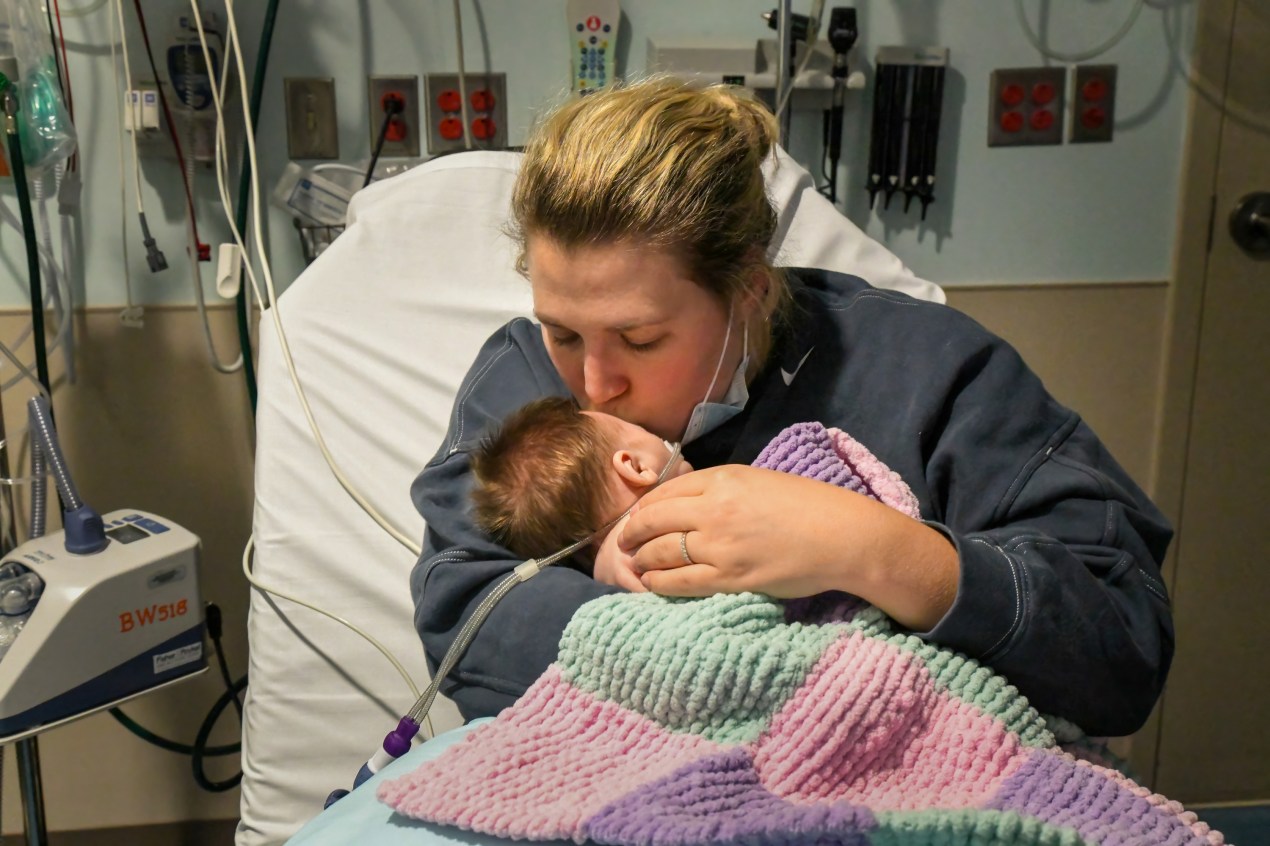 Caitlyn Houston, who wears a grey sweatshirt and has her blonde hair pulled up in a loose bun, holds her infant daughter in her arms while kissing her on the forehead. They are sitting in an emergency room.