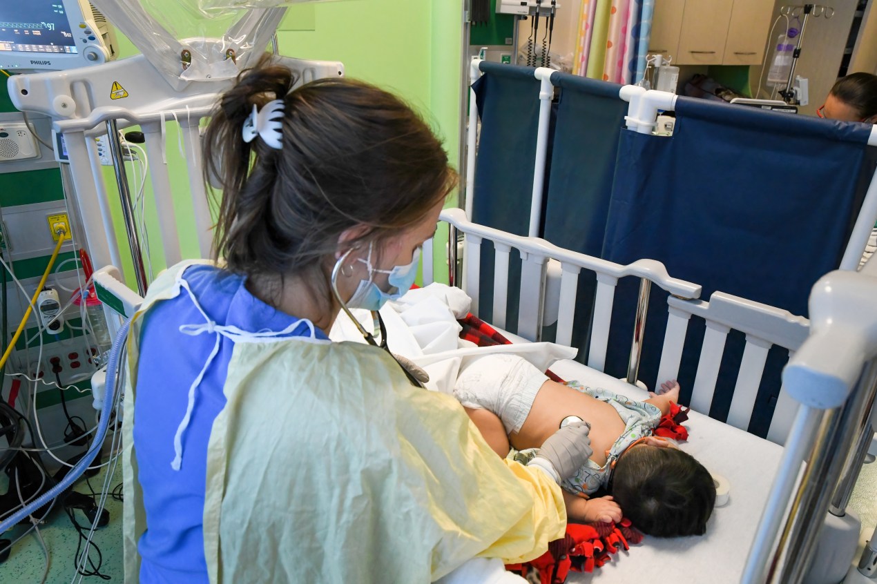 A hospital worker is caring for a newborn baby in the pediatric care unit, which resembles a crib.  The infant lay on his stomach with his head turned away from the camera. 