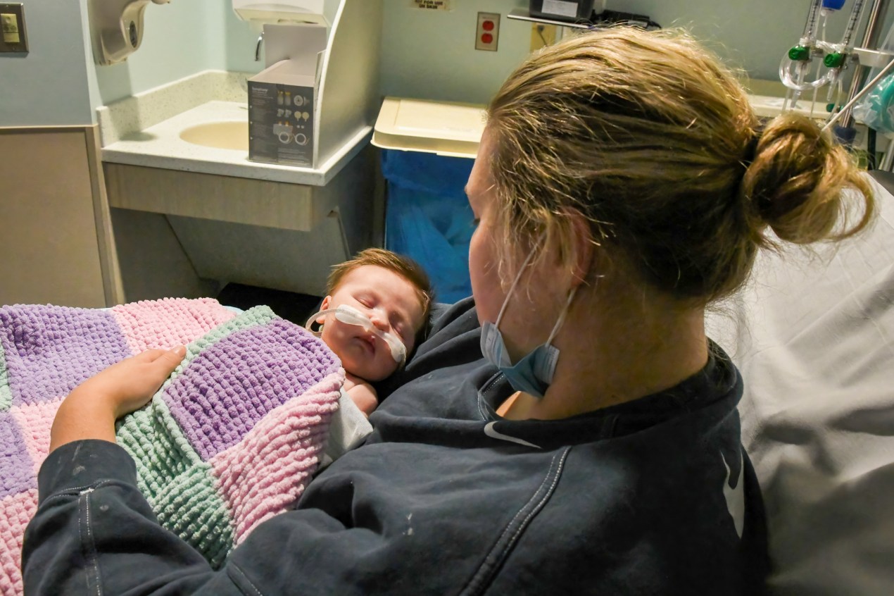 Caitlyn Houston holds her infant daughter, Parker, as they wait in the emergency department.  The photo was taken from above the mother's shoulder, revealing the infant's face.  The newborn baby has an oxygen tube on his face, under his nose.  She is sleeping.