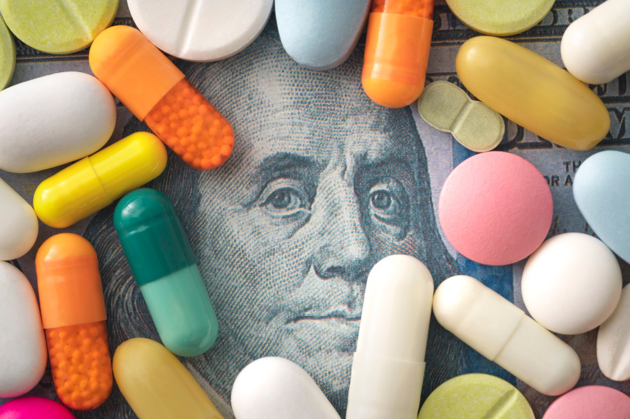 Various pills of different colors and shapes cover a 100 dollar bill. In the center, the face of Benjamin Franklin is visible.