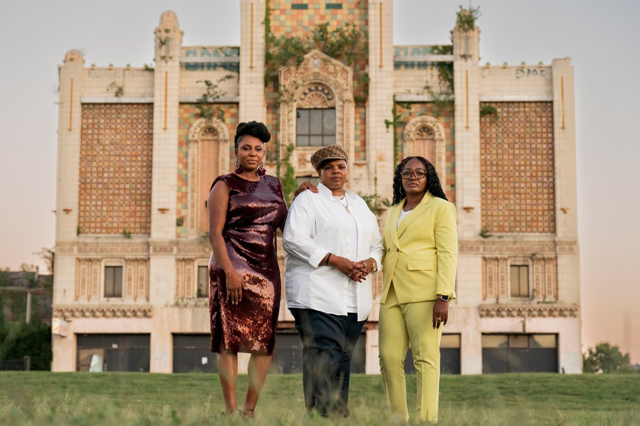 A photo shows three Black women posing for a portrait together outside at sunset. They stand in front of East St. Louis' Majestic Theater, a large Gothic building.