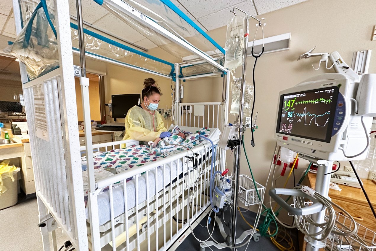 A photo shows a nurse with a stethoscope checking on an infant inside a hospital intensive care unit.