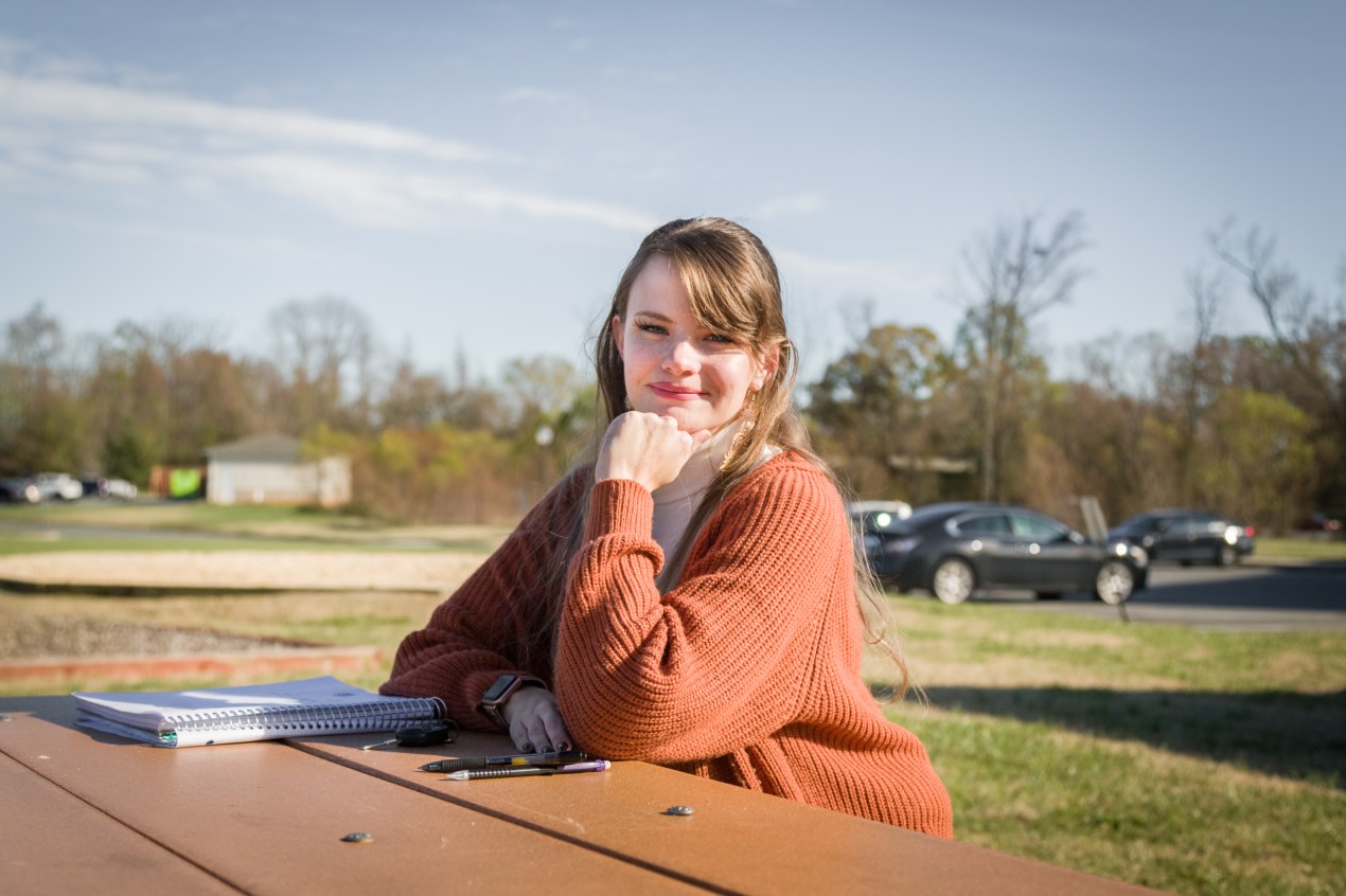 Lauren Overman, who has strawberry blonde hair with bangs and wears a loose orange sweater, is sitting outside at a picnic table. Green grass and blue sky are visible behind her. She looks directly at the camera with a smile.