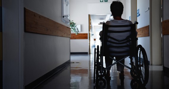 A photo shows a senior woman sitting in a wheelchair in a nursing home corridor.