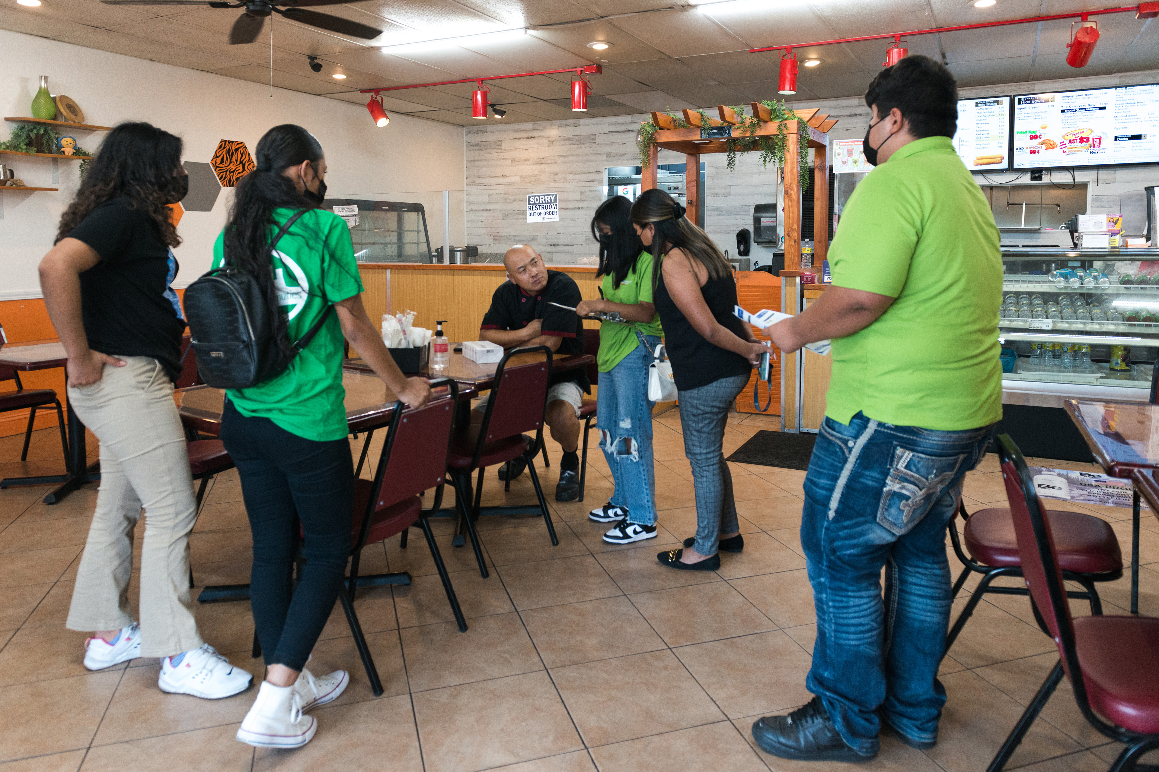 Teens deliver covid tests and information flyers to Chris Vang, a restaurant owner. Vang is seated at a table inside the restaurant while the teens stand around him.
