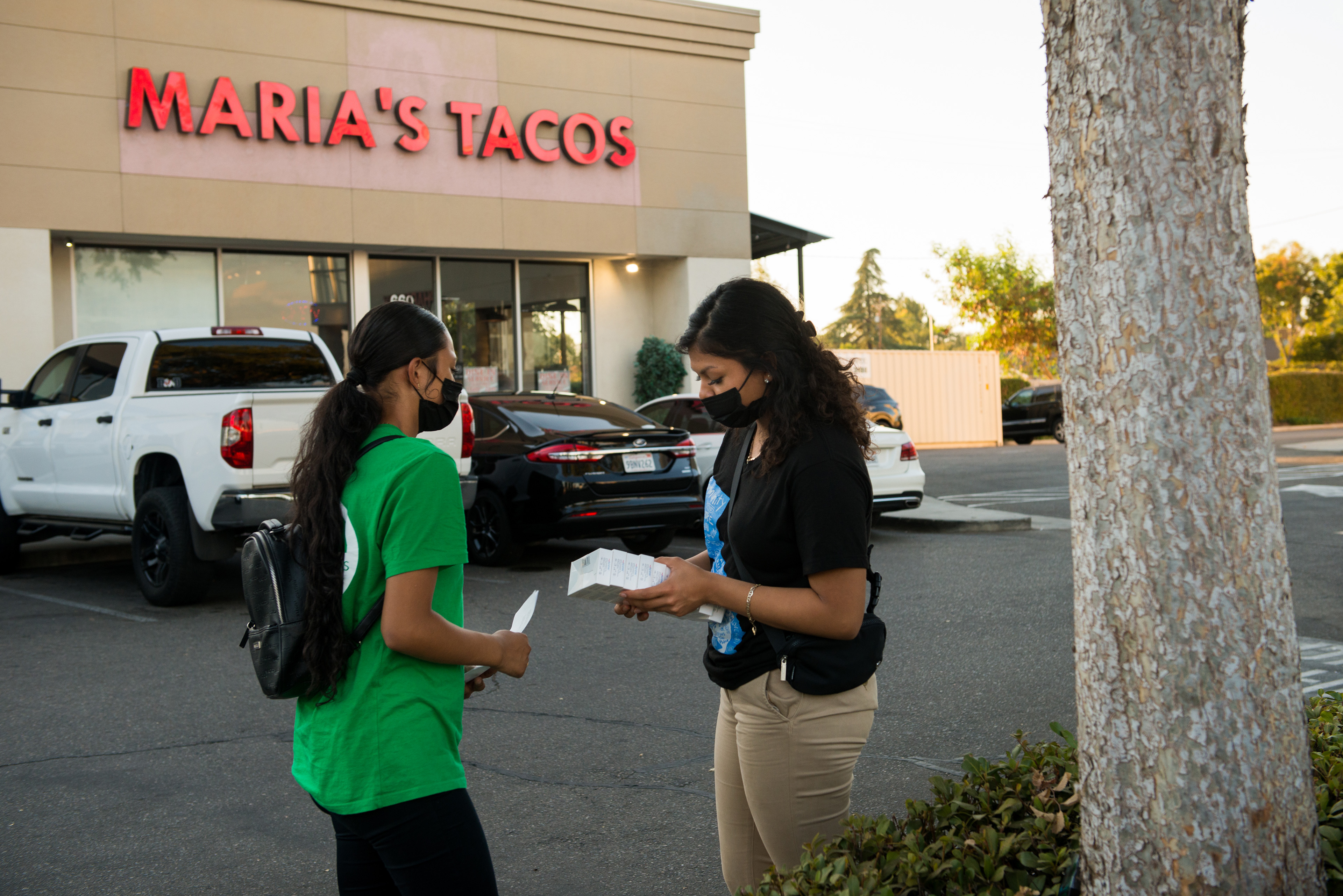 Melissa Lopez (solda) ve Alma Gallegos (sağda) covid testi dağıtmaya hazırlanıyor.  yazan bir binanın önündeki otoparkta duruyorlar. "Maria'nın Tacoları."