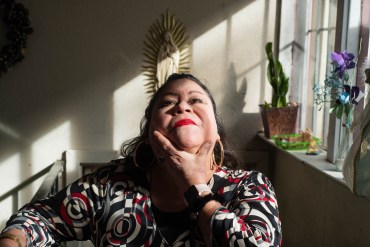 Bertha Embriz is sitting on a chair in her house beside the window. The sun is shining in, highlighting her face as she looks up to the ceiling. In the background, a statue of the Virgin Mary can be seen hanging on the wall, as well as some plants on the windowsill.