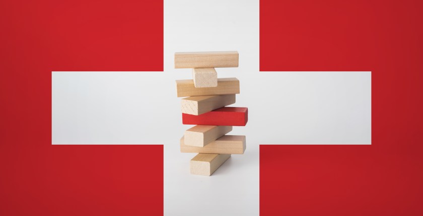 A stack of wooden blocks is seen surrounded by a medical cross. One of the wooden blocks in the center of the stack is painted red, indicating the tower may fall.