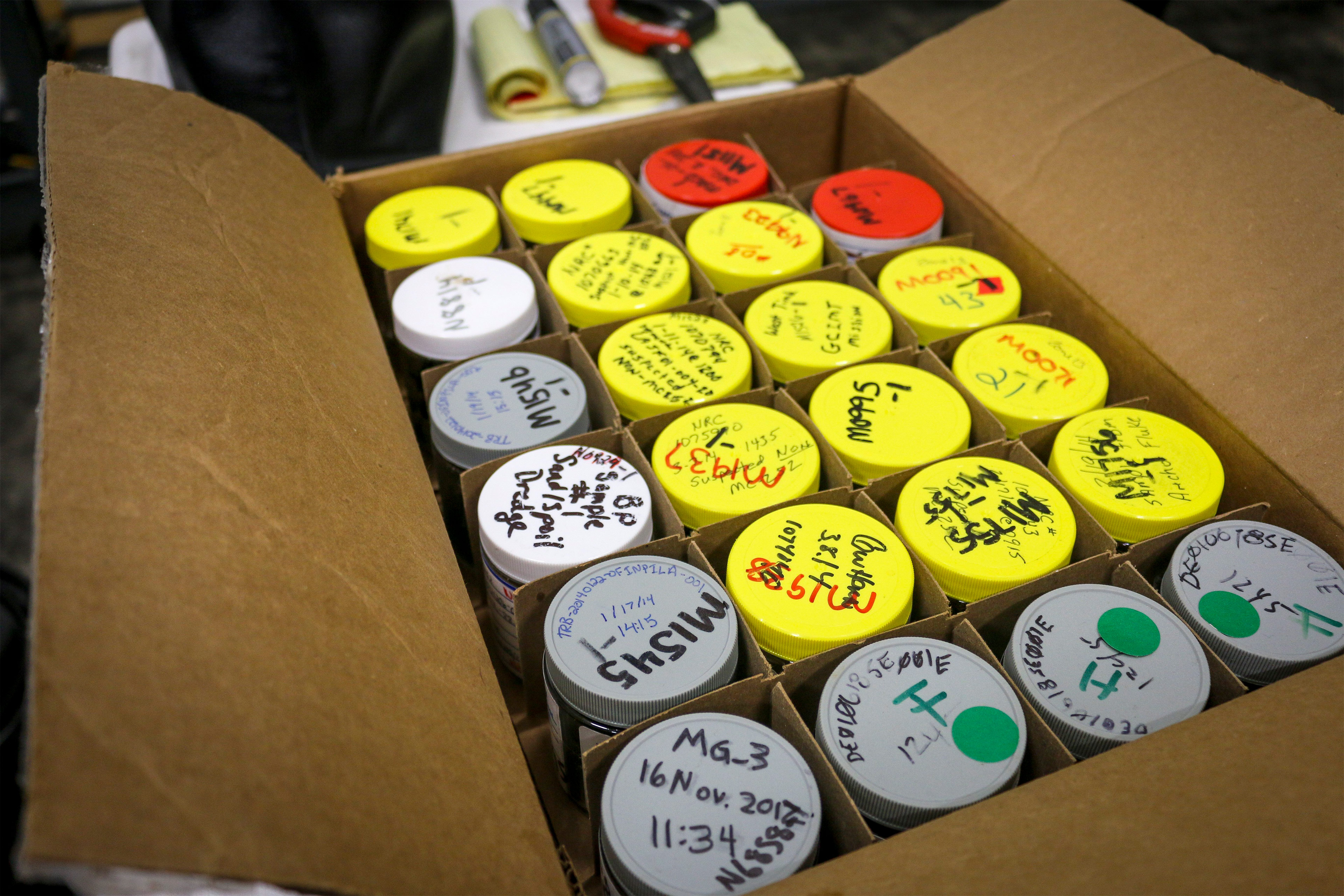 A photo shows a cardboard box filled with jars with handwritten labels on top of them.