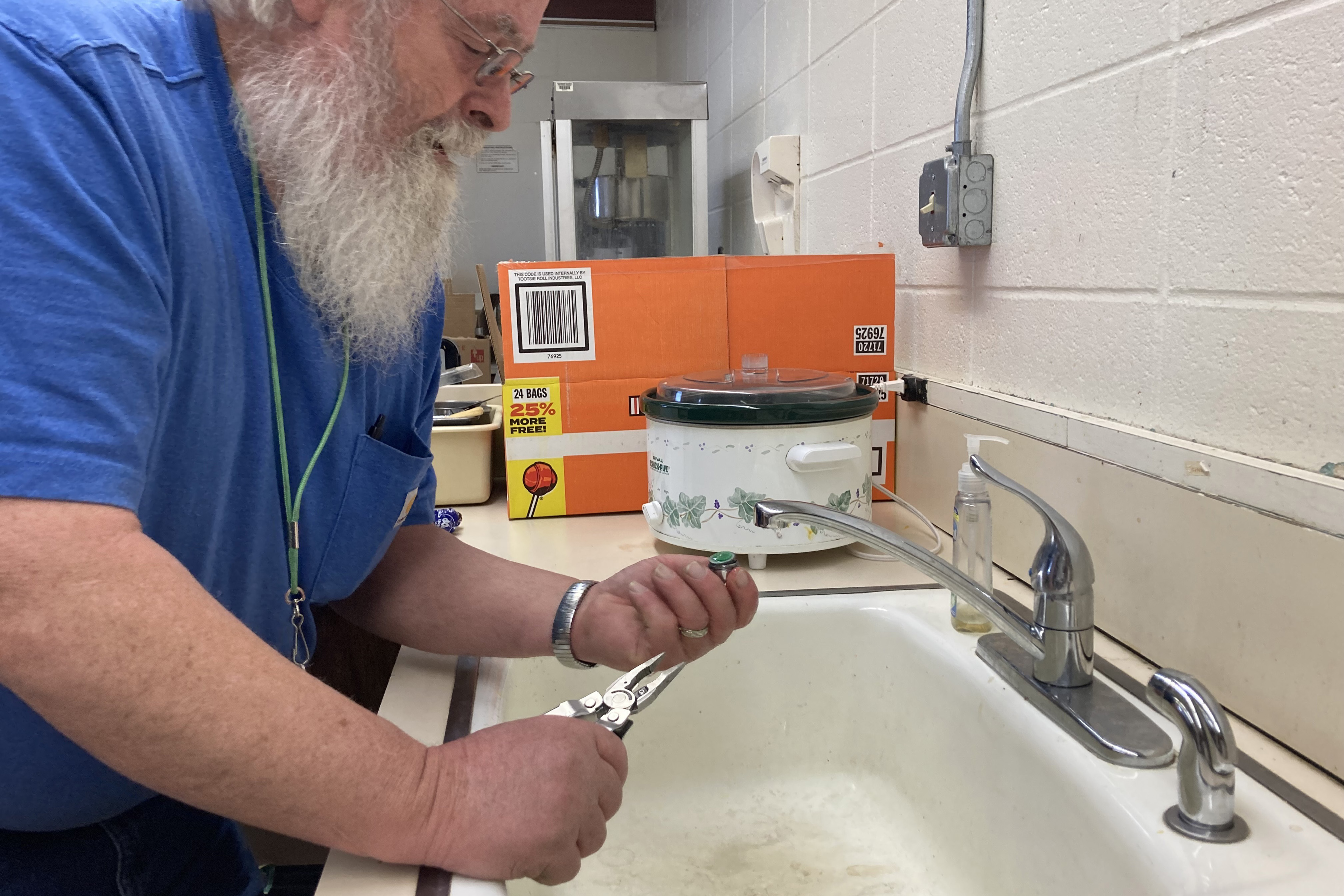 Chris Cornelius is leaning over a sink examining a faucet which he has removed. In his other hand, he holds a wrench.