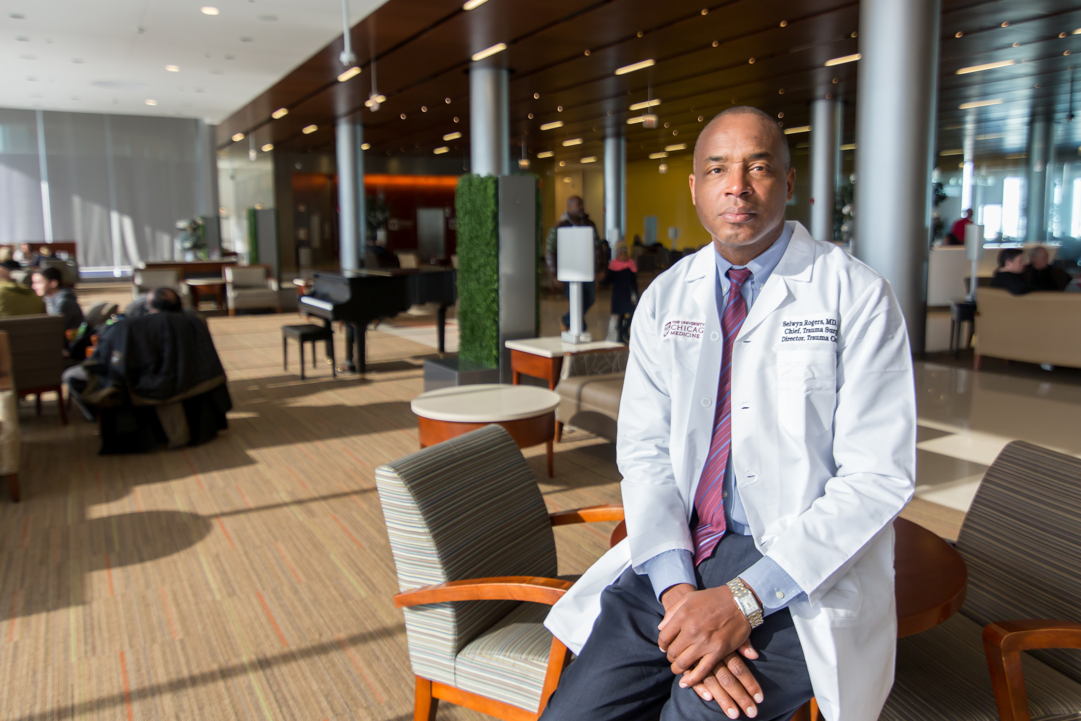 Dr. Selwyn Rogers sits on a chair in a hospital lobby. He wears a white doctor's coat and looks directly at the camera. The room is sunny and spacious.