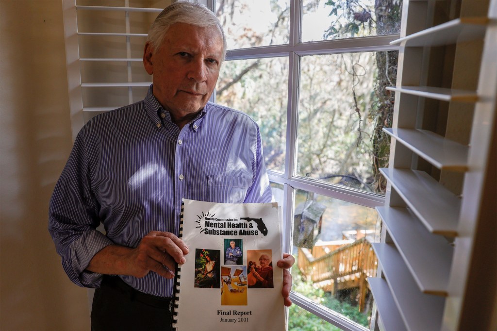 A photo of a man by a window holding a copy of Florida's 1999 mental health commission report.