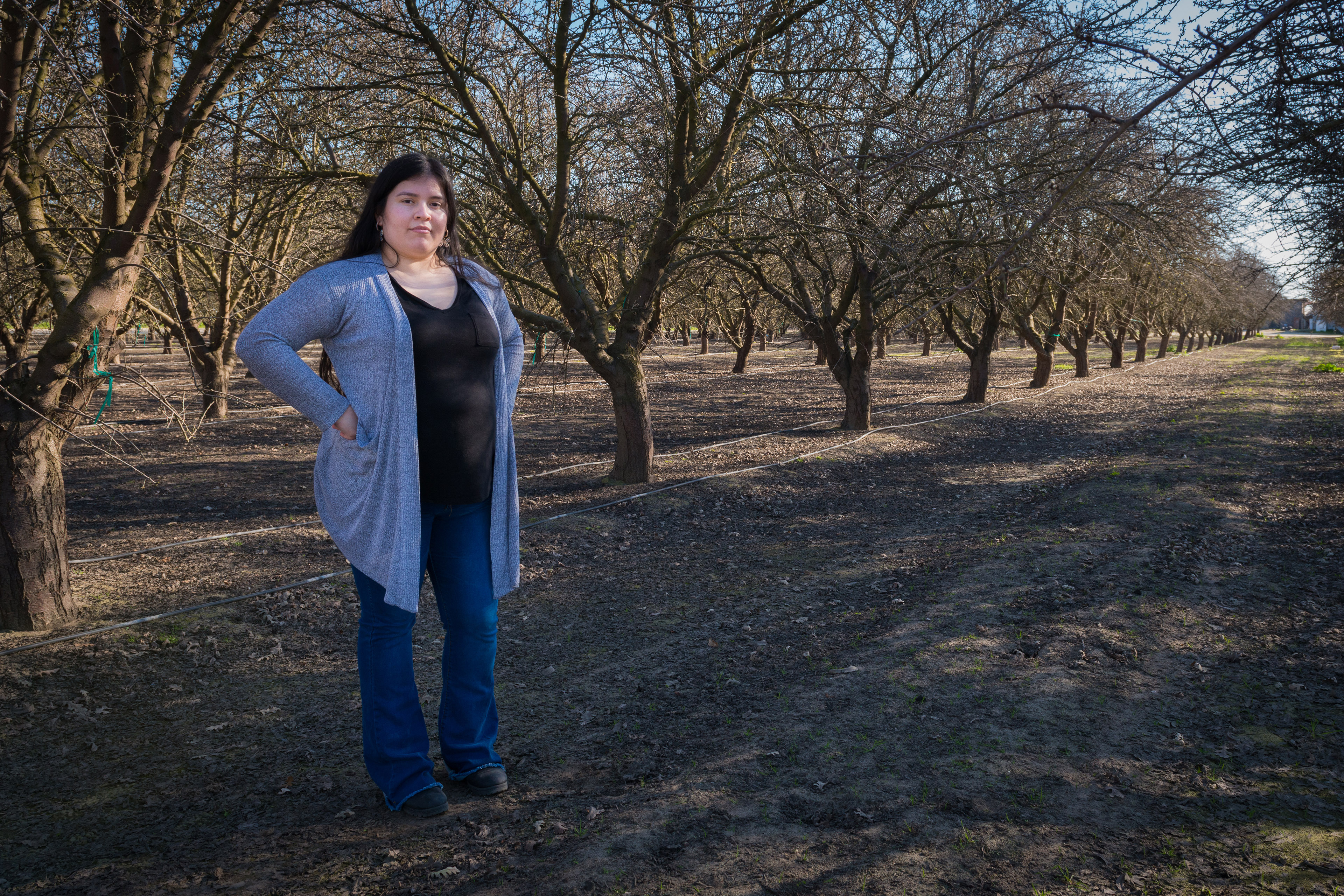 Carolina Morga Tapia stands outside, amid almond trees. The trees have no leaves, signaling the time of year.